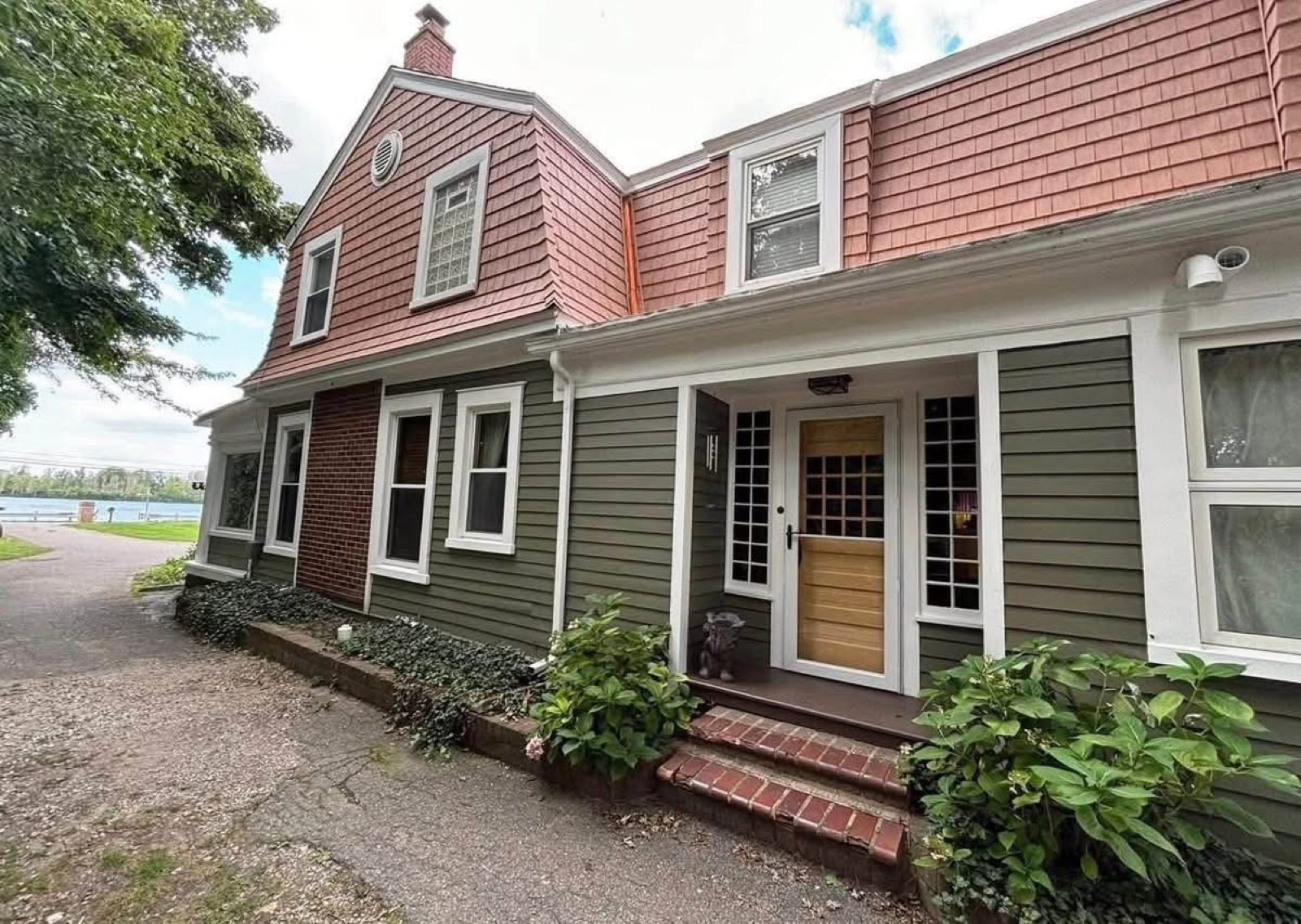 Waterfront Dutch Colonial home with green lap siding, a red shingled gambrel roof, brick accents, and a natural wood front door