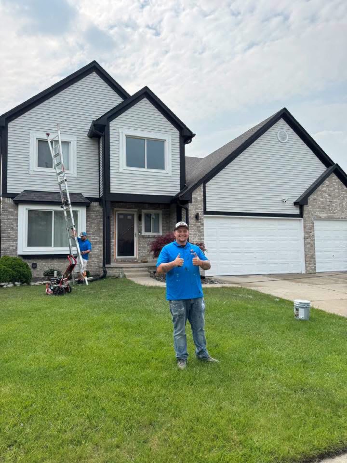 Large two-story home with gray lap siding, black-trimmed windows, and a white garage door, with a Brake painter in the front yard mid-project