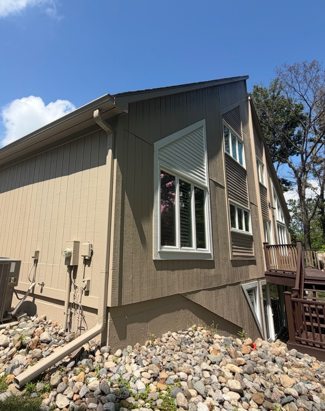 Two-tone exterior repaint with taupe lower siding and darker olive board-and-batten upper walls, white-trimmed windows, and a side deck
